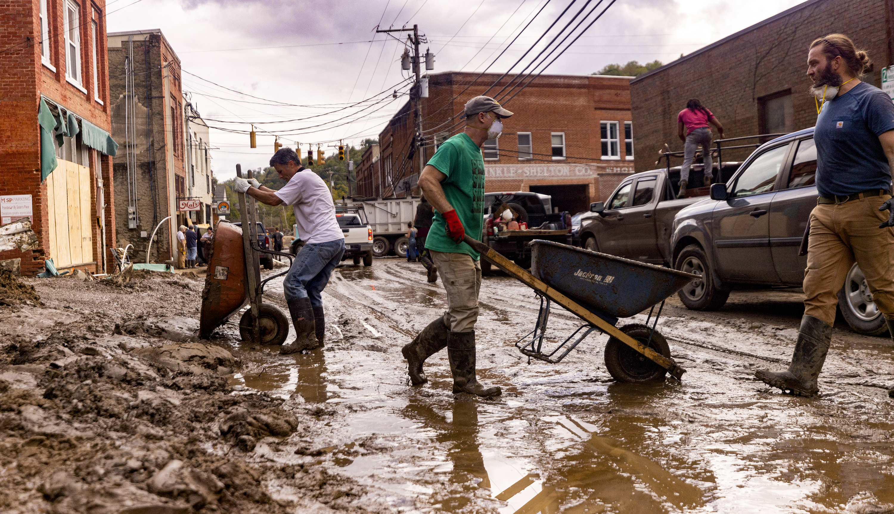 Men in boots push wheelbarrows full of mud through a flooded street.