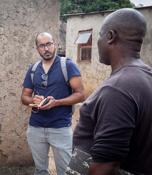 Photo showing a reporter with a notepad interviewing a man standing amid a cluster of rural buildings.