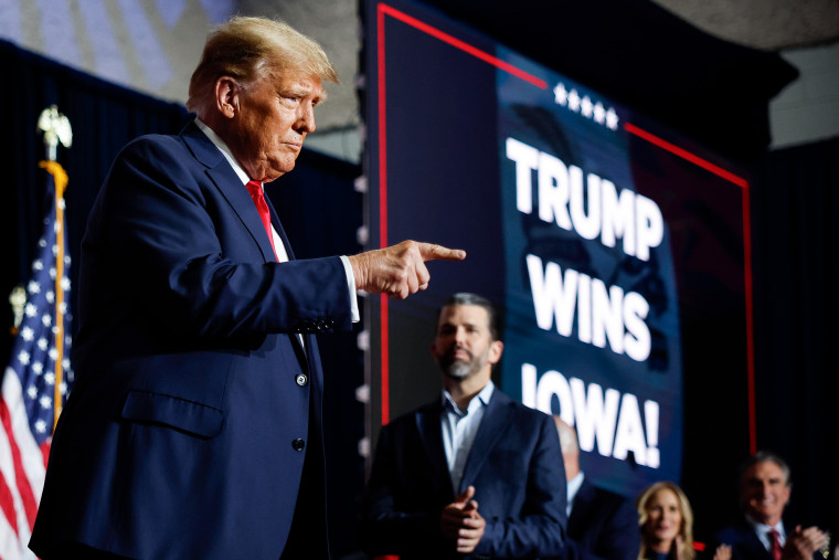 Donald Trump points next to a projected screen that says "Trump wins Iowa"