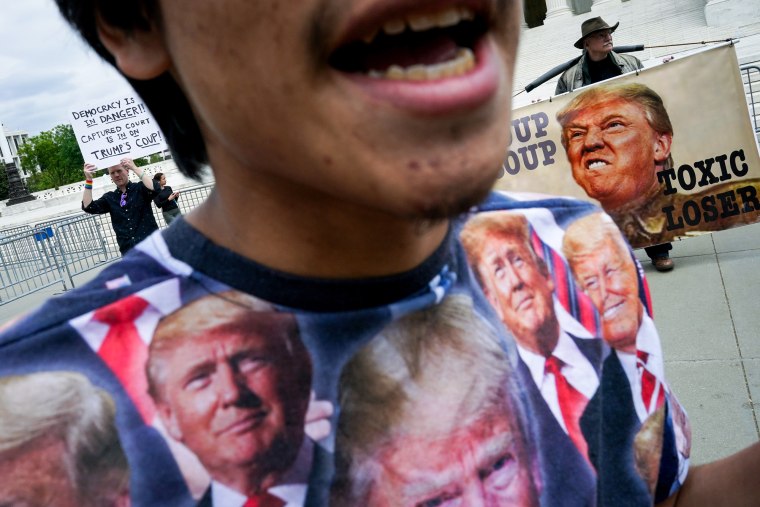 A man wearing a T-shirt with various Trump faces in front of anti-Trump protestors with posters in the background, outside