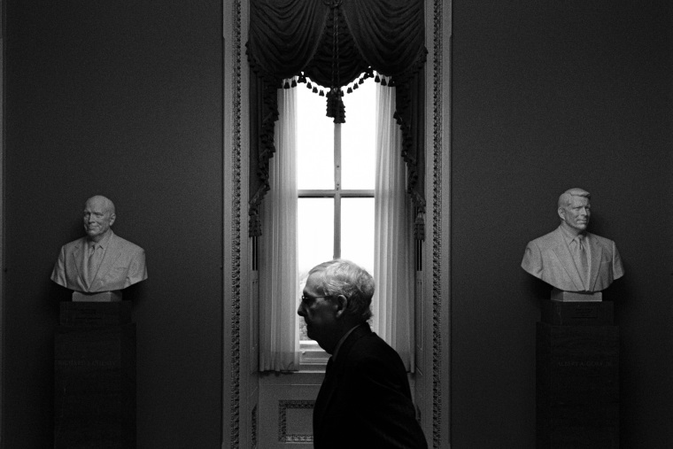 Mitch McConnell walks indoors, seen between two busts in a hallway while passing by a window