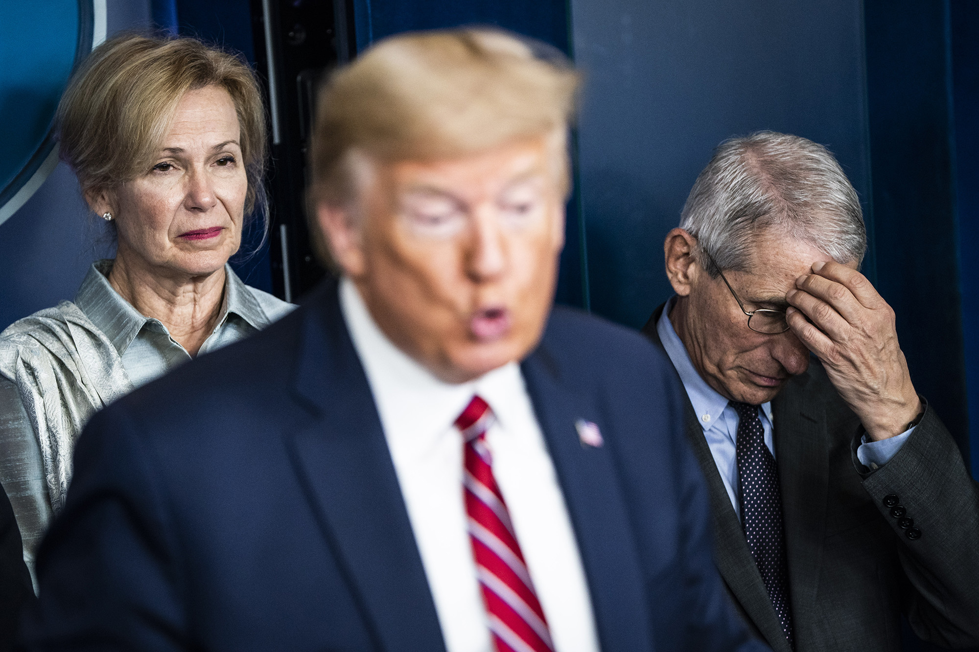 Donald Trump out of focus in the foreground with Dr. Deborah Birx behind him to the left and Dr. Anthony Fauci to his right, with his head in his hand.