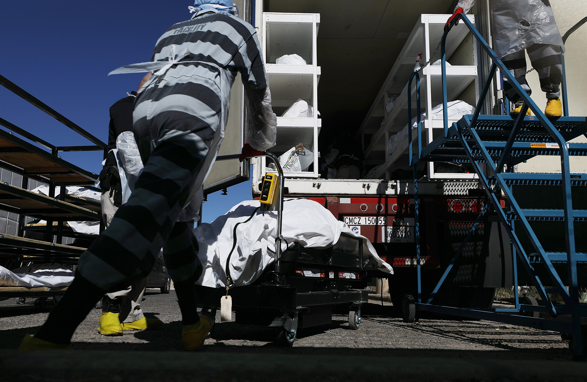 Low angle photo of a man in a black and white striped jail uniform pushing a cart with a dead body on it to a cargo truck stacked with other bodies.