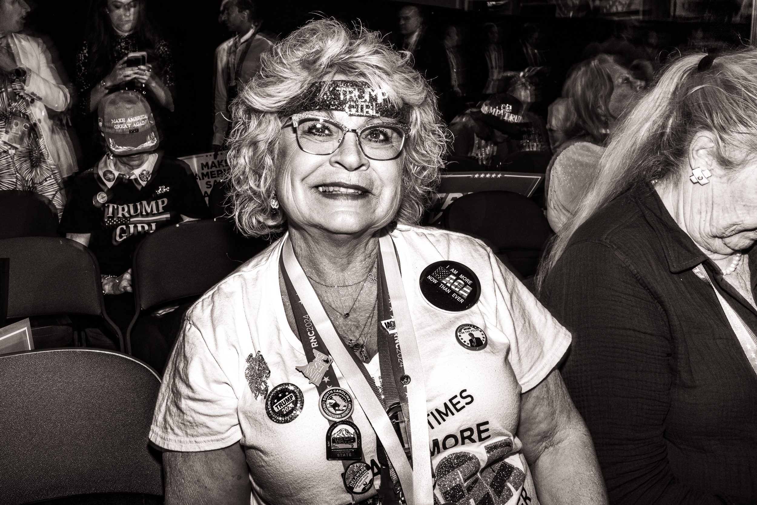 Black-and-white photo of a woman smiling. She's wearing a glitter headband that says, "Trump Girl."