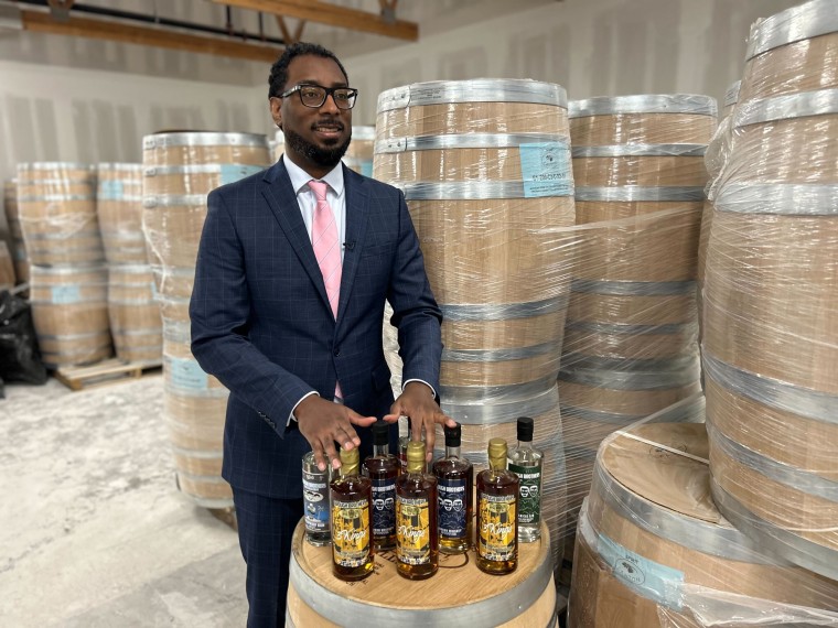 Victor Yarbrough stands near barrels and places his hands atop bottles of liquor, posed for a photograph