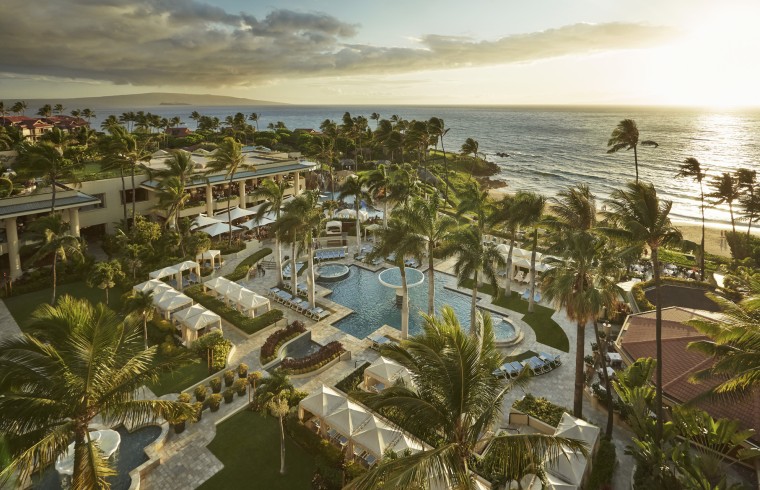 An aerial view of a hotel resort, palm trees, cabanas, a pool, and the ocean can be seen