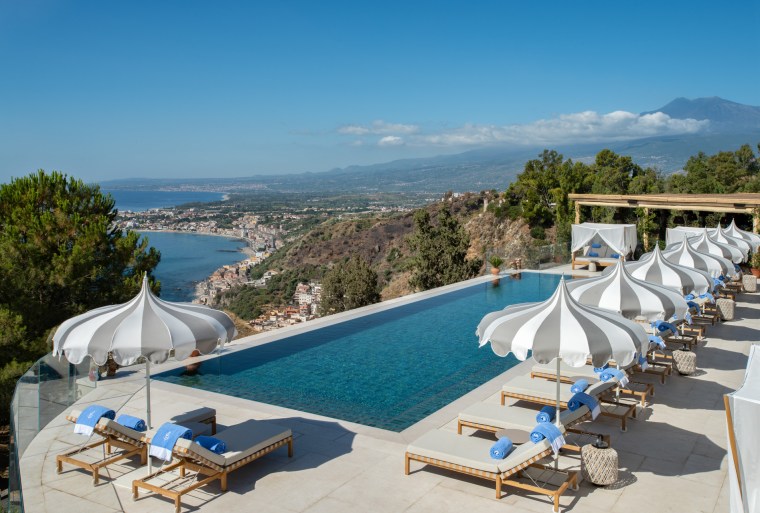 A view of the ocean and hillsides is seen outdoors from a pool surrounded by poolchairs and umbrellas