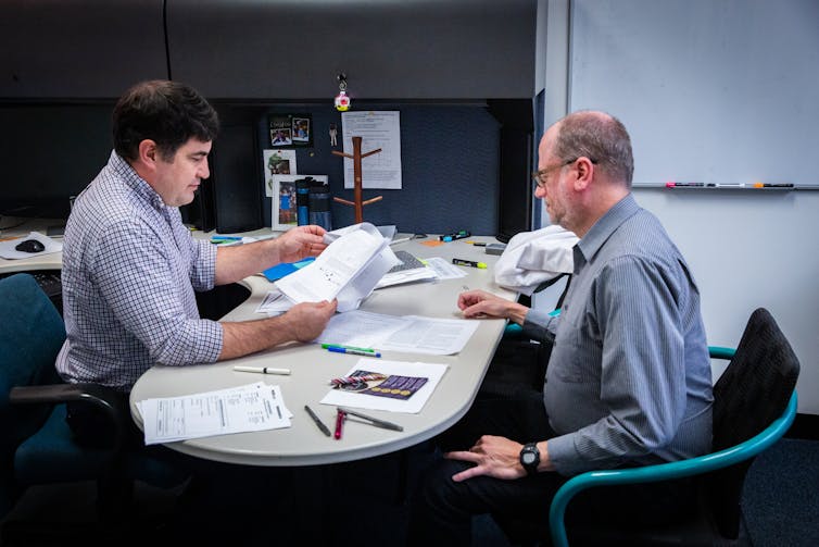 Two men sitting across from each other at a table filled with papers